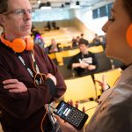 A young woman in an LED lit wearable tech jacked and headphones talks to an event visitor about Ling Tan’s project SUPERGESTURES