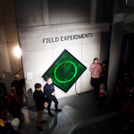 Looking down on a stage from above, three men are speaking into a microphone in front of a green sign that says Field Experiments