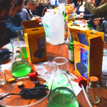 A group of people sit around a long table, all looking at a woman speaking and holding a laptop. A science jar full of green liquid is on the table