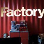 Red stacked boxes appear in front of a red metal wall, above in gold is the word 'Factory'