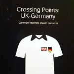 A computer illustration of a white t-shirt against a black background, and the words Crossing Points: UK-Germany in white font