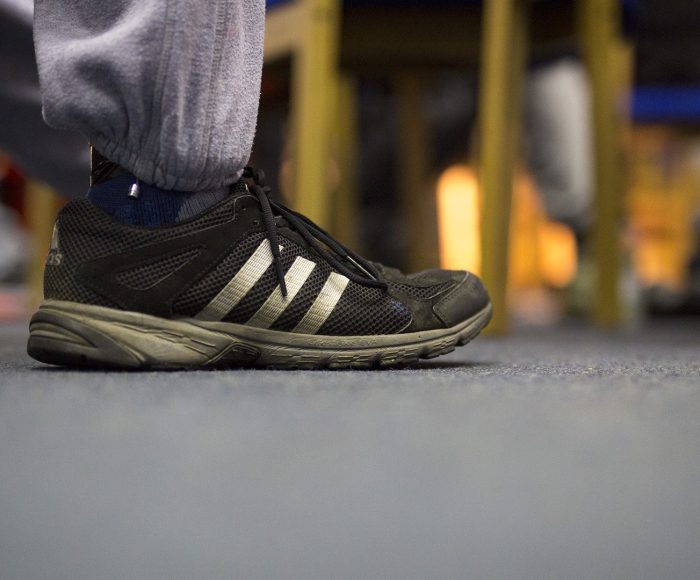A man's foot placed on a grey carpet, he is wearing grey jogging pants and black adidas trainers covered in mud