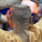 Man in prison overalls is seated facing forward with his hands raised in the air, taking part in an auction